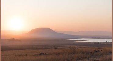 A photograph capturing the sunset over the Ntshingwayo (chelmsford) Dam. Also in the photo is a reddish/ pink sky, a little mountain, grasslands and the dam.