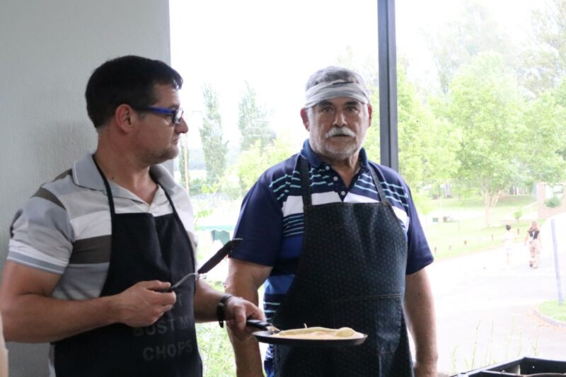 Two men are standing by a window. The older man has a bandanna over his forehead. He is looking straight at the camera. The younger man, is wearing glasses and holding a pan with pancake in it. He is holding a spatchela in his right hand. Both are wearing aprons.