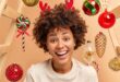 Close up shot of curly haired woman smiles broadly has white teeth and healthy dark skin wears red reindeer antlers looks gladfully at camera expresses happiness surrounded by Christmas toys