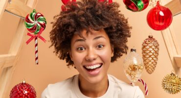 Close up shot of curly haired woman smiles broadly has white teeth and healthy dark skin wears red reindeer antlers looks gladfully at camera expresses happiness surrounded by Christmas toys