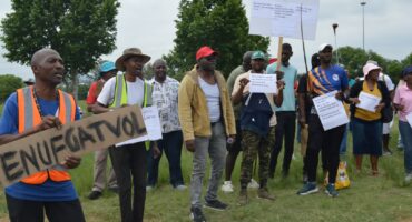 Protestors dressed in colourful attire standing on a grassy field with trees in the background, holding up various placards demanding rates and taxes be reassessed.