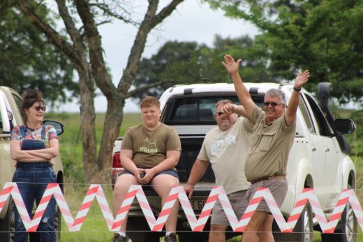 Image of two men, a teenage boy and a woman waving at the camera while standing on the other side of a drag race track.
