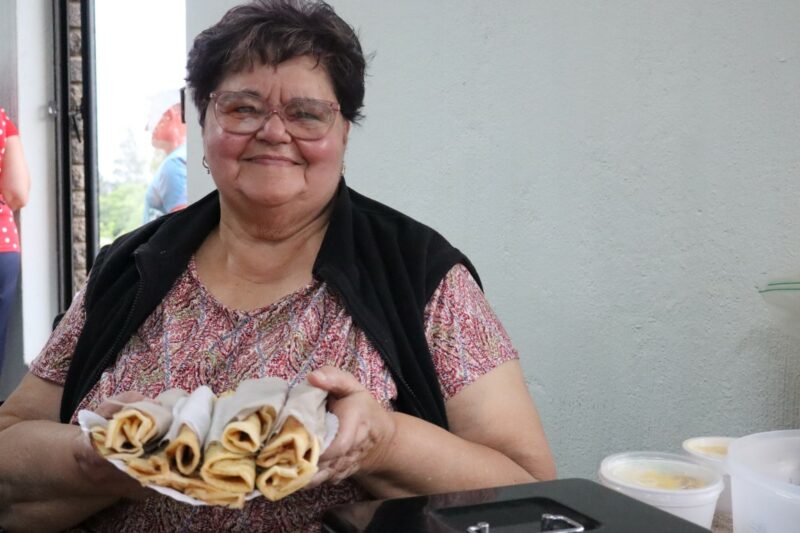 A lady with red cheeks and glasses is holding a paper plate full of rolled up pancakes. She is indoors. She is wearing pink and black. She has short, dark hair.