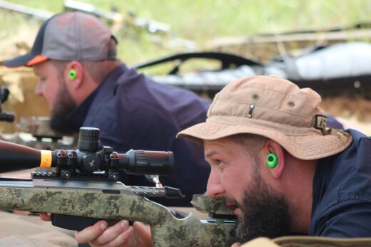 Two men lying on the ground on their stomach, with firearms in their hands, aiming at the gongs. One man with a bucket hat and other with a cap.