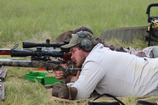 Two men lying on their stomachs with firearms, aiming at the gong, one with cap and headgear.
