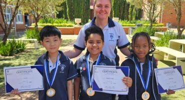 One grown-up and three children are photographed holding certificates, outside in front of a school building.
