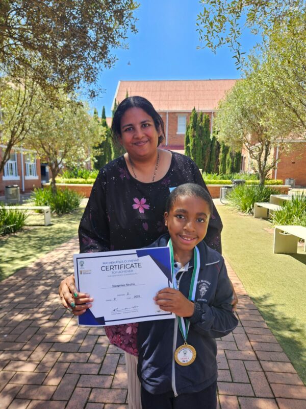 A teacher and her learners hold up certificates outside in front of their school building.