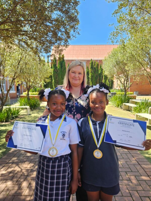 A teacher and her learners hold up certificates outside in front of their school building.