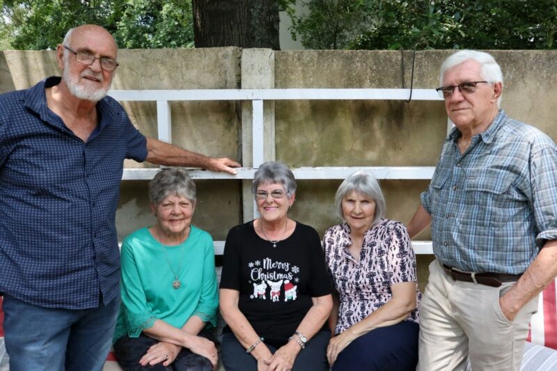 Five people posing for a photo in front of a concrete wall. There is white wooden planks against the wall. Three ladies are sitting, two men are standing. The people are elderly. It is outdoors.