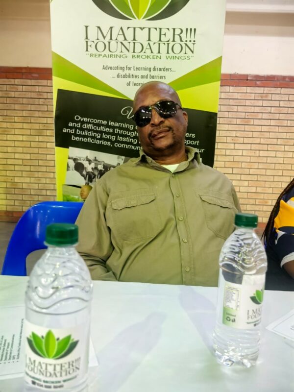 A man wearing sunglasses sits at a panel table in front of an I Matter Foundation banner, with two branded water bottles placed on the table.
