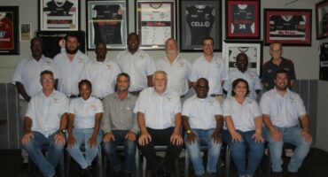Several men pose for a formal photo infornt of a series of rugby related framed photographs.