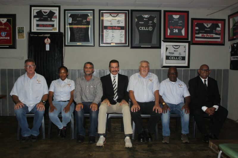 Several people pose for a formal photo infornt of a series of rugby related framed photographs.