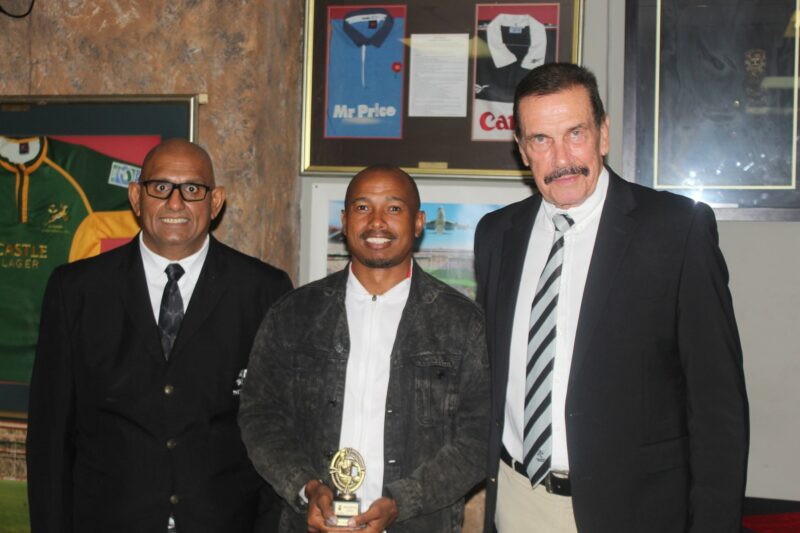 Three people pose for a formal photo while receiving a trophy, standing in front of a wall with rugby memorabilia on the wall.