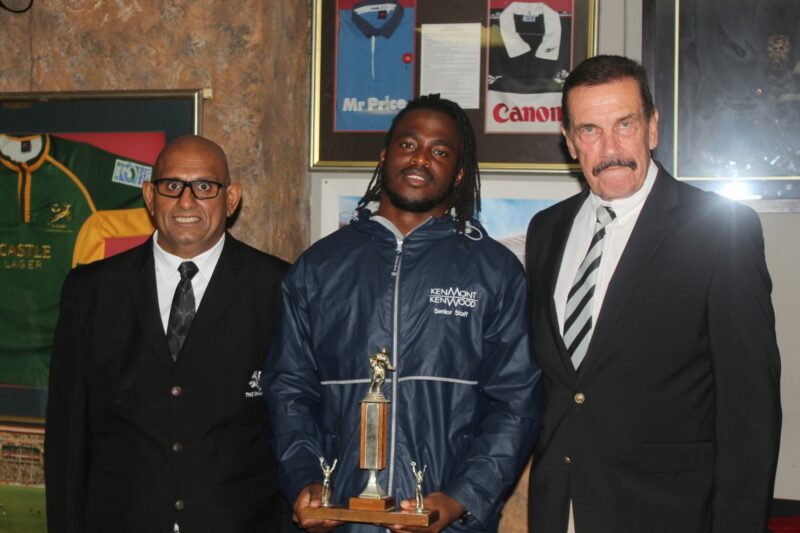 Three people pose for a formal photo while receiving a trophy, standing in front of a wall with rugby memorabilia on the wall.
