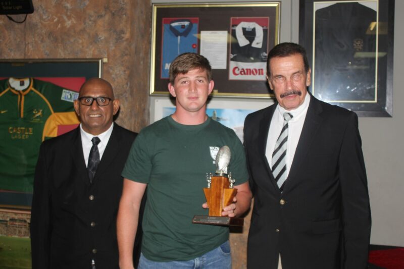 Three people pose for a formal photo while receiving a trophy, standing in front of a wall with rugby memorabilia on the wall.