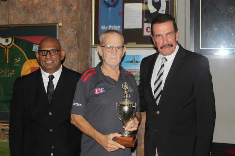 Three people pose for a formal photo while receiving a trophy, standing in front of a wall with rugby memorabilia on the wall.