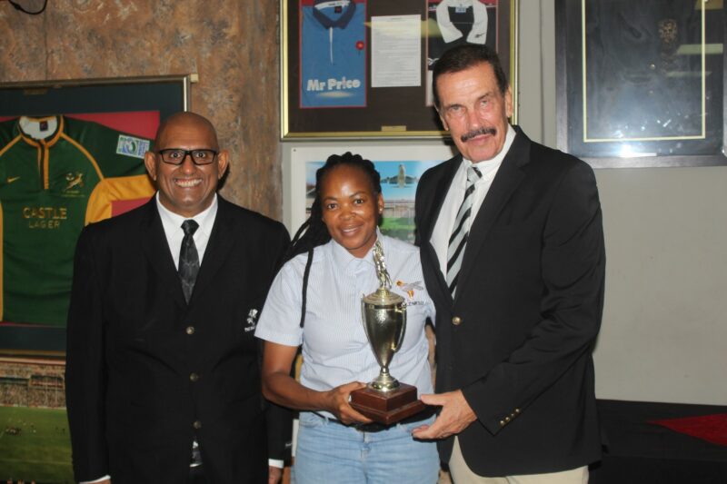 Three people pose for a formal photo while receiving a trophy, standing in front of a wall with rugby memorabilia on the wall.