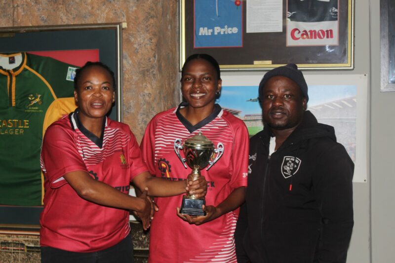 Three people dressed in rugby attire posing for a photo with a trophy in front of a wall full of rugby memorabilia.