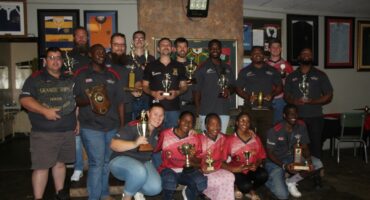 Several Rugby Players pose for a photo inside a rugby club holding their trophies in front of a wall with rugby memorabilia.