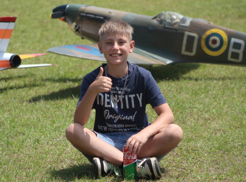 A young boy shows a thumbs up while posing in front of two model aircrafts.
