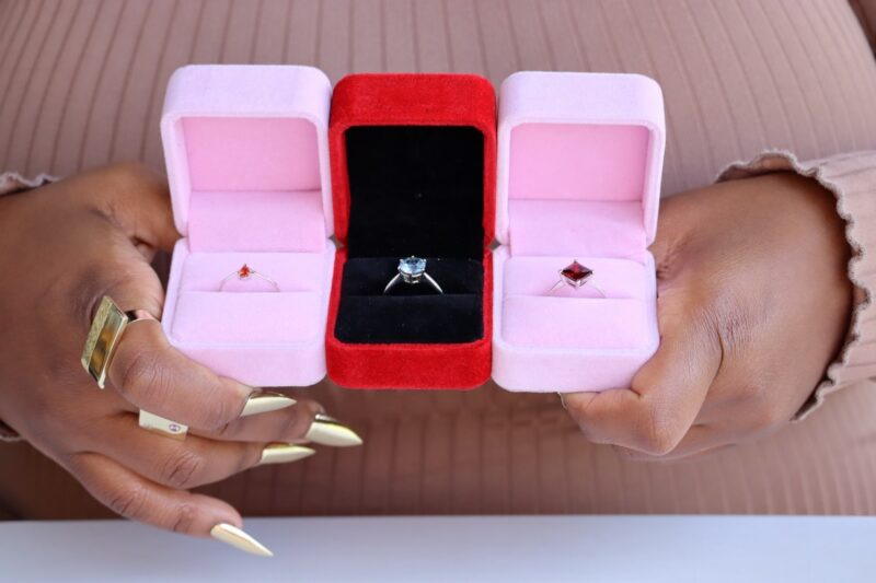 Three ring boxes. Tow pink, one red. There are rings inside the boxes. A lady with long, brass nails are holding the boxes in front of her chest.