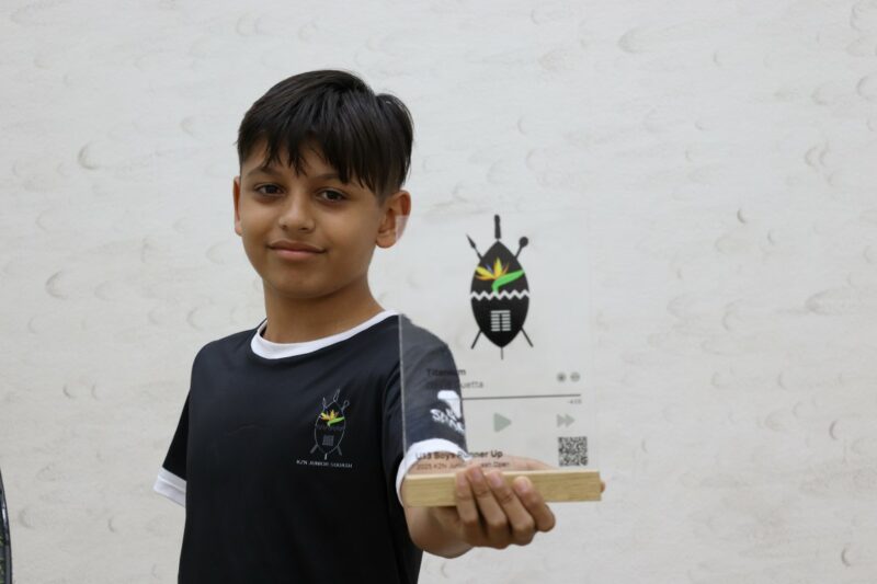 A young boy with black hair and black clothing is holding a transparent, glass trophy in his left hand.There is a shield on the trophy with writing on it.