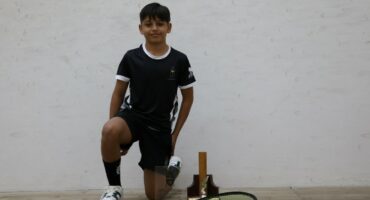 A young boy is kneeling in front of a white wall. The floor is made of wood. There are two trophees and a squash raquette in front of him. He is wearing black, and his shoes are white.