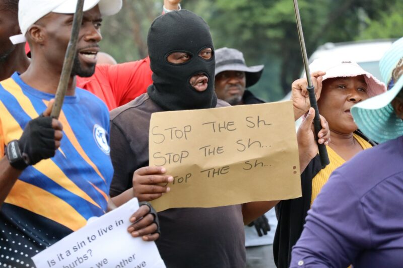 A protestor is wearing a black mask. He is holding a placard in his right hand. There are many people around his. They are wearing different colours.