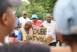 A poster with "Dube" and "Gatvol" written in balk. A man with a blue shirt is holding the placard. There are people behind the man haolding the placard.