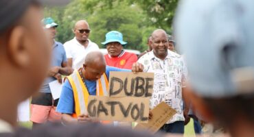 A poster with "Dube" and "Gatvol" written in balk. A man with a blue shirt is holding the placard. There are people behind the man haolding the placard.