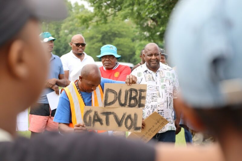A black man is wearing a blue shirt and a visability vest. He is holding a placard with the words "Gatvol Dube" written on it. He is surrounden by other people. The man with the placard is looking down.
