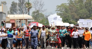 A large group of people are marching down a city street. They are holding placards in their hands. THere is police officer in front of the crowd. A man is holding a placard with the word "gatvol" written on it. It is during the day, there weather is hot.