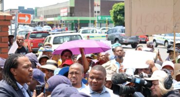 A group of people protest outdoors, holding placards with messages to the Newcastle municipality. Public Order Police Officers stand in infron of the protestors, busy protecting the building behind them.