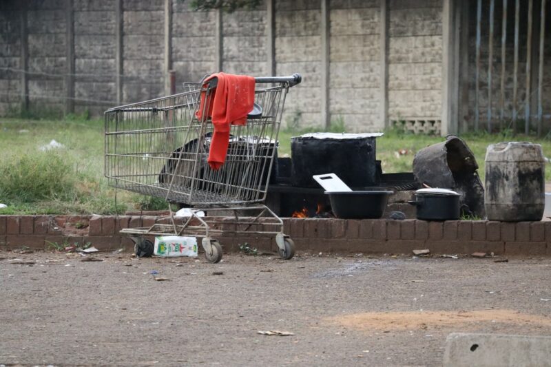 A shopping trolley in a parking lot with a red sweater draped over it. There is a white packet stuck under the trolley. Behind the trolley is an open fire with materials to make food. The materials are black with a white cutting board. There is a concrete wall in the background.