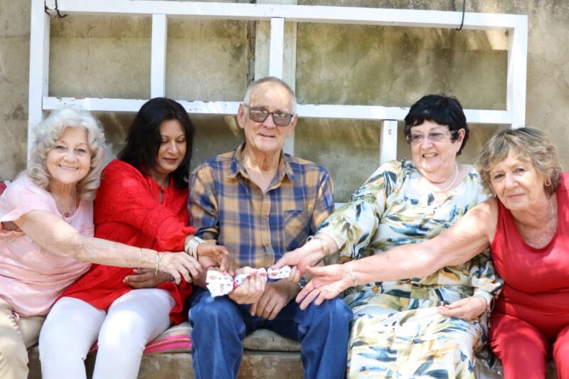 Four ladies are sitting both sides of an elderly man. He is holding a white item in his hand. The ladies are all grabbing the cracker in his hands. They are sitting with white wooden planks in the background.They are outdoors.