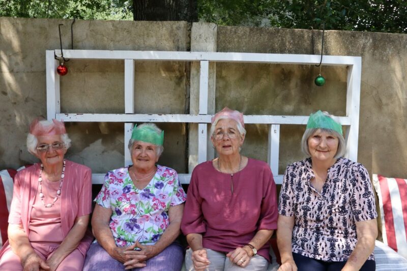 Four elderly ladies are sitting in front of a concrete wall. There are white, wooden plans against the wall. All four ladies have paper crowns on their heads. They are wearing various shades of pink. They are outdoors.
