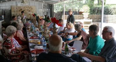 A group of retirees sitting around a table. The table is decorated with Christmas decorations. There are plates, cuttlery and drinks on the table. The table is under a shed-like roof. It is day-time.