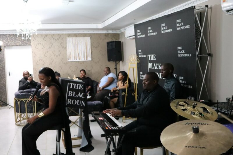 A band of four people. There is a singer, guitarists, keyboard player, and drummber. They are all dressed in black. The singer is sitting in front of the band. There is a group of people sitting in the corner in the background. There is a black banner with white writing against the wall. It is a well lit room.