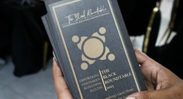 The Black Roundtable's program for the evening's event. It is black and gold, with writing in gold. A female with long nails is holding the program. There is a picture of a round table with four figures sitting around the table. The floor and some black chairs are in the background.