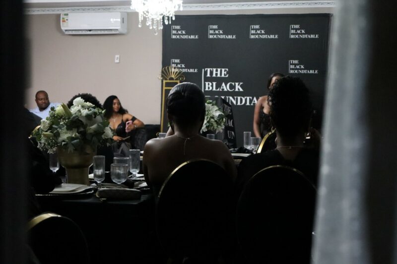 A photo of guests from behind. They are looking at a female singer in front of the room. There is a black banner with white wording gainst the wall. Some guests are sitting on black and gold chairs, others are sitting in the corner on black sofas.