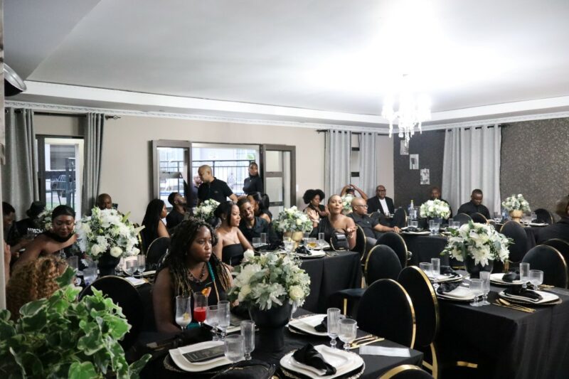 It is a well lit room. There a decorated tables with people sitting around it. The people are all doing their own thing. The tables and people are dressed in black and gold. There is flower arrangement on each table. There are curtains in front of the windows. The door at the back of the room is open.