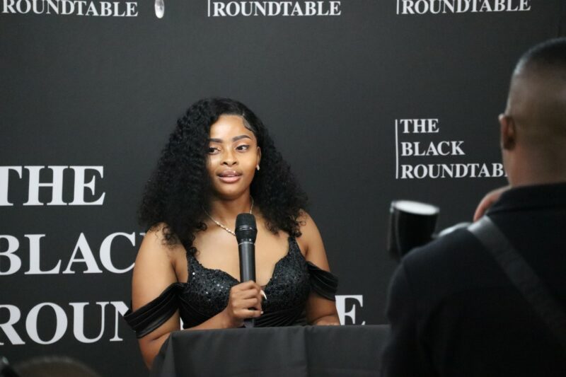 A woman is standing behind a podium. She is speaking into a black microphone. The microphone is in her right hand. She is wearing black, her hair is shoulder-length. There is a camera-man in the foreground with a camera in his hand. There is a black banner with white wording behind the lady.