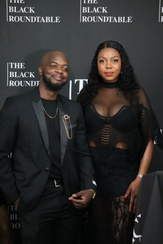 A man and a woman dressed in black in standing in front of a black banner with white wording. The man has a gold jewellery piece on the left side of his jacket. The woman is looking straight in front of her and the man is looking slightly to the left. The man is smiling, the woman is not.