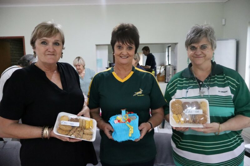 Three ladies are standing with baked goods in their hands. Two of them are wearing green, one is wearing black. There is a kitchen behind them.