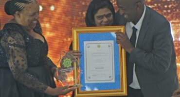 Three people are pictured celebrating an award during an award ceremony. A woman is seen holding a trophy while smiling at another woman holding a framed certificate, with a man also in the picture.