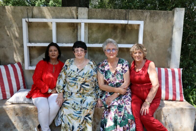Four Ladies are sitting in front of a concrete wall. There are white, wooden planks on the wall. Two ladies are wearing dresses, two ladies are wearing pants. The ladies are wearing white, read, green, pink and flowers. They are outdoors.