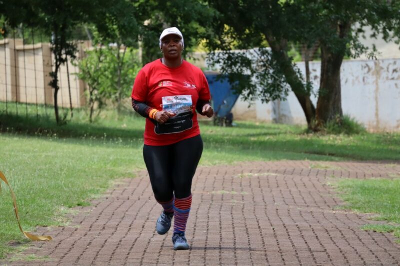 A woman is running. She is wearing red shirt with long stiped socks and a black pants. Her shoes are blue. She has something on her head. She has a bag strapped around her waist.She is running on cobblestones in the outdoors.
