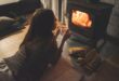 A woman lies on the floor near a wood-burning stove while holding a mug and watching the fire.