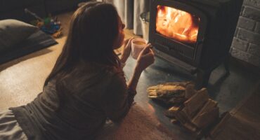 A woman lies on the floor near a wood-burning stove while holding a mug and watching the fire.