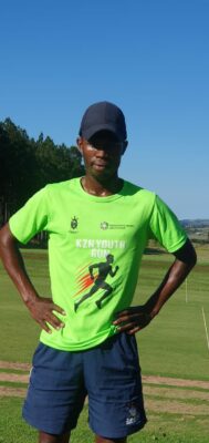 African man standing on parkrun course with green shirt and blue shorts and hands in his sides.